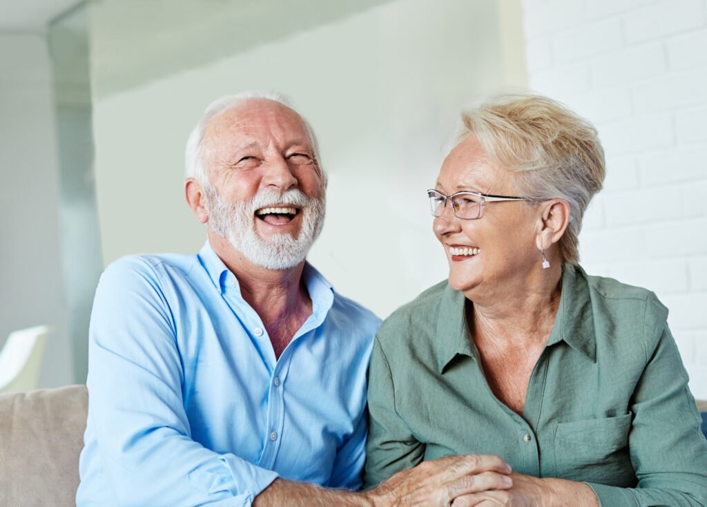 An elderly couple is sitting together, laughing joyfully.