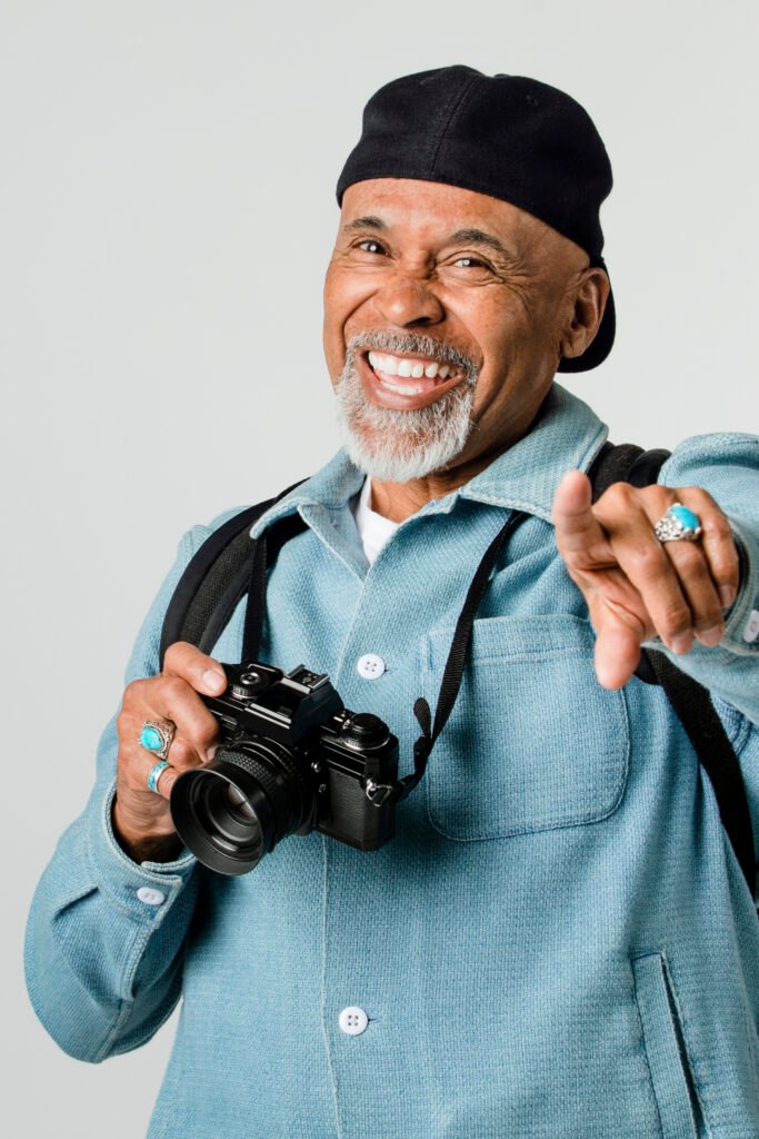 A smiling man in a blue jacket points playfully while holding a camera.