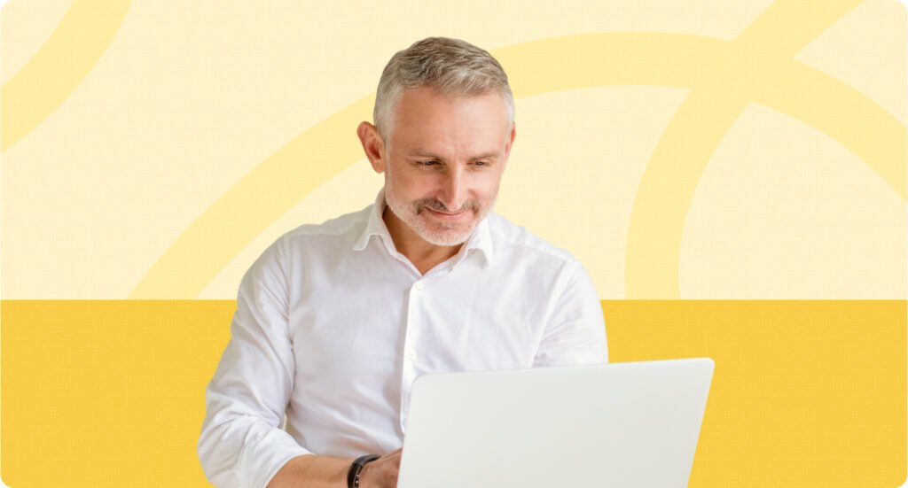 A gray-haired man in a white shirt looks at a laptop.