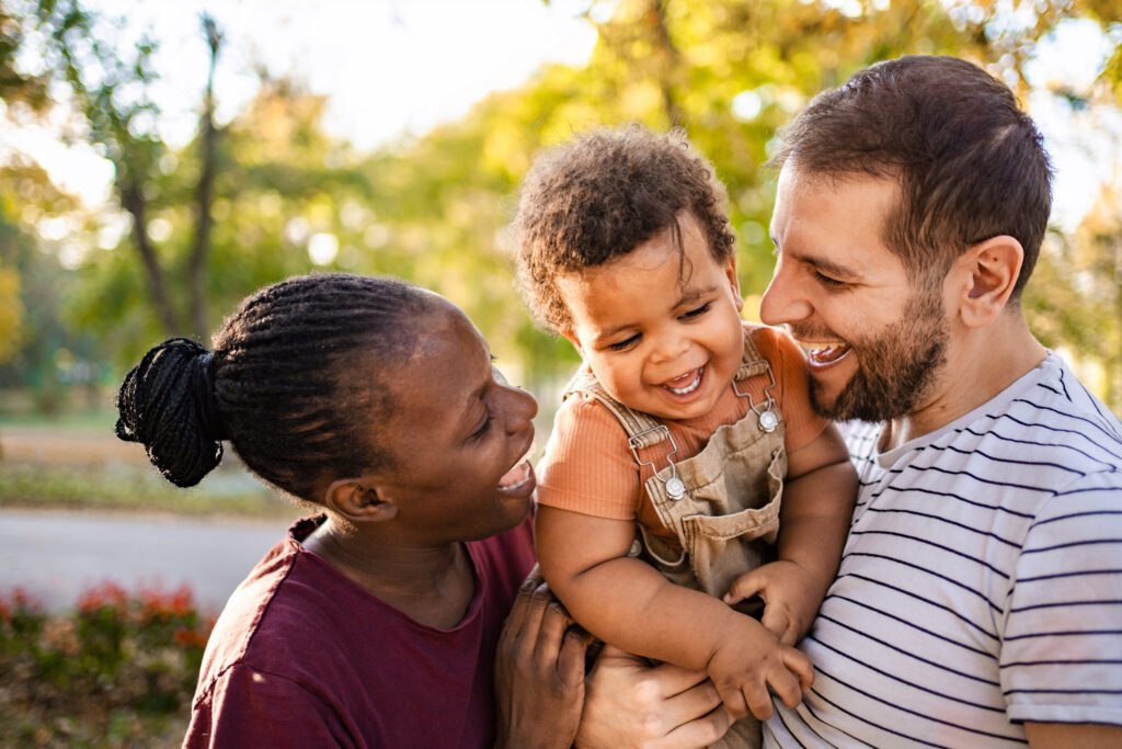 A loving couple enjoys a delightful day in a vibrant park, laughing together as they embrace their joyful child. The golden sunlight filters through the trees, enhancing their happiness.