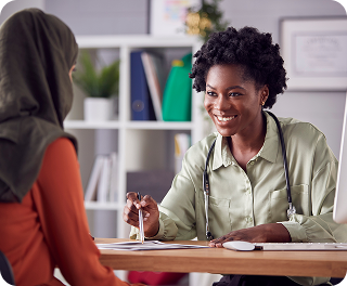 A doctor with a stethoscope smiles and points at a document while talking with a patient wearing a headscarf.