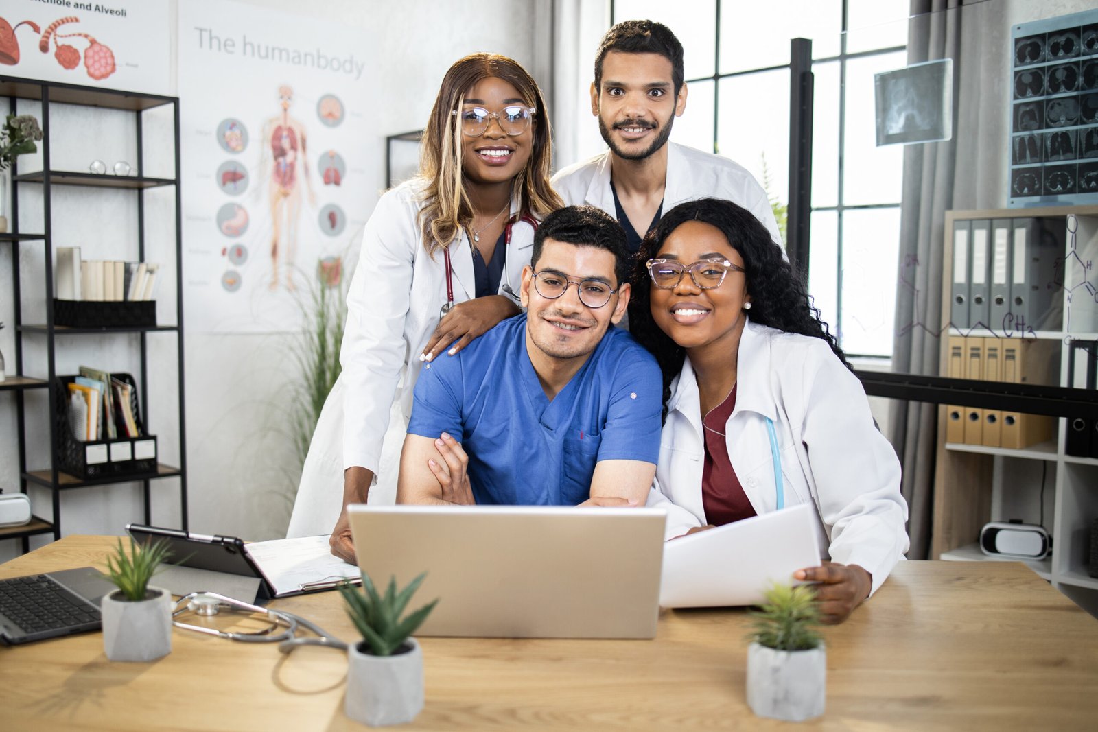 Group of four international doctors in lab coat and scrubs sitting together at table and using laptop and tablet. Medical conference of male and female specialists. Sharing with knowledges.