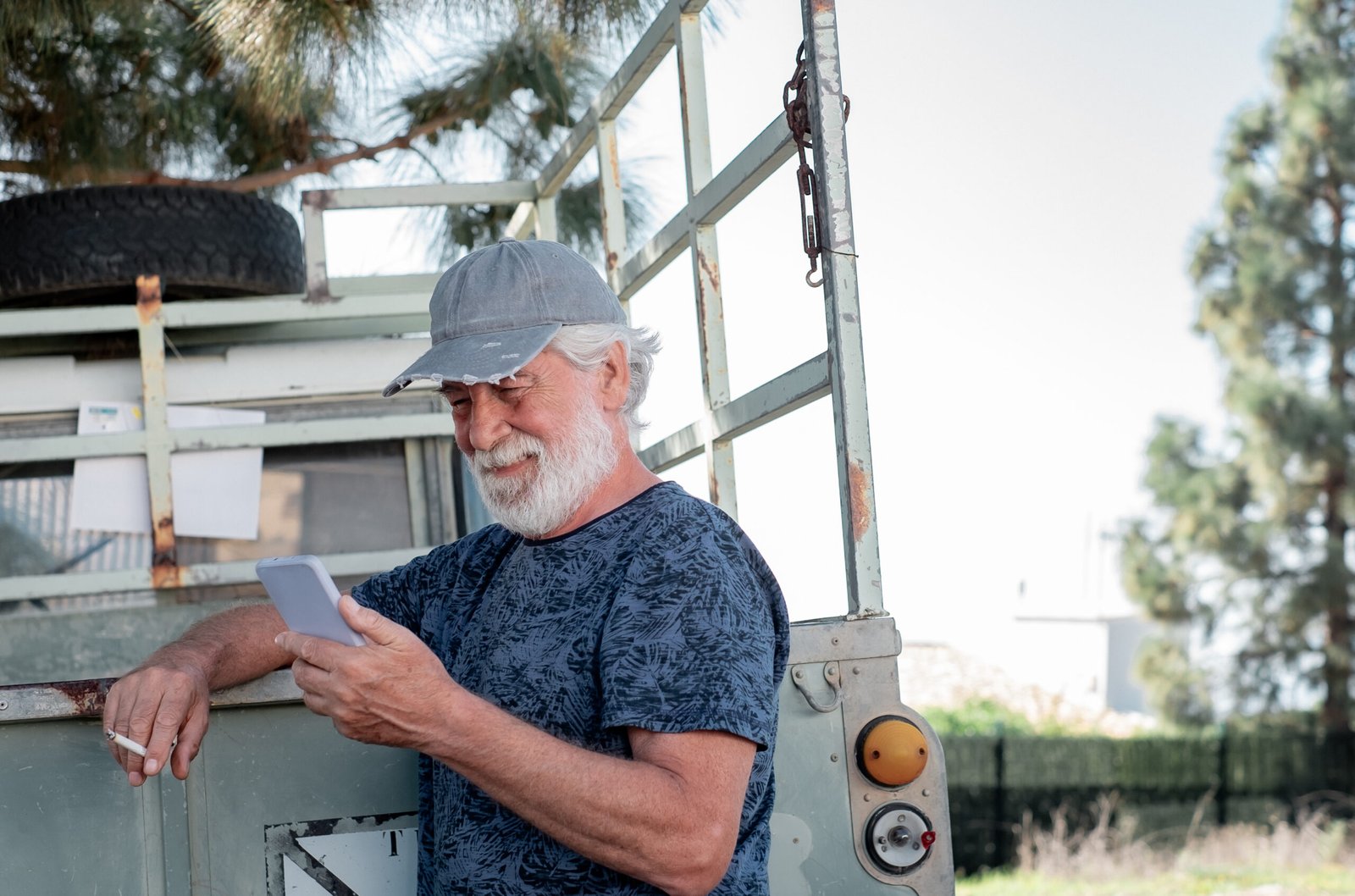 Senior white haired bearded man with cap leaning against his van smoking cigarette and holding mobile phone